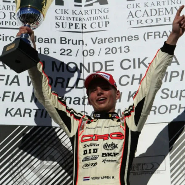 A race driver in a cream and black racing suit with red trim and a red cap raises a trophy above his head, standing in front of a white backdrop printed with event text and sponsor logos.