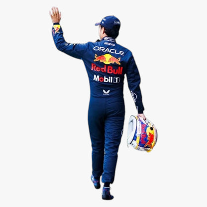 A Red Bull Racing driver seen from behind, wearing a dark blue Oracle Red Bull Motorsport suit with sponsor logos, a matching cap, and carrying a colorful Red Bull helmet against a plain white background.