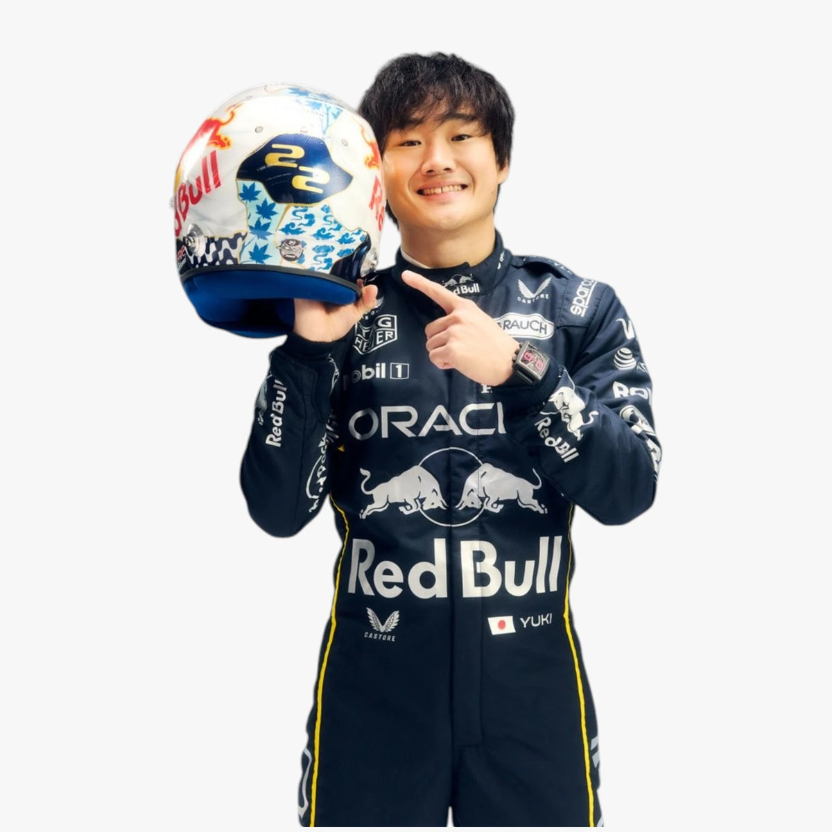 A smiling Red Bull Racing driver in a dark navy team suit holds a white, blue, and red decorated racing helmet while pointing at it, against a plain light gray background.
