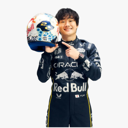 A smiling Red Bull Racing driver in a dark navy team suit holds a white, blue, and red decorated racing helmet while pointing at it, against a plain light gray background.