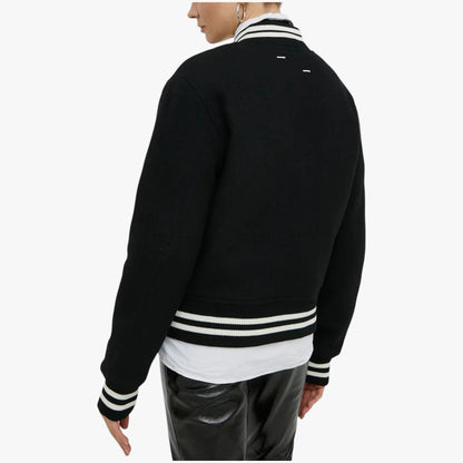 A person shown from the back wearing a black varsity-style jacket with white striped collar, cuffs, and hem, layered over a white shirt and black pants, against a plain light gray studio background.