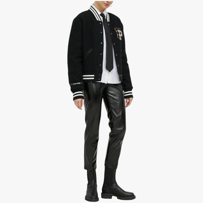 A model wearing a black varsity jacket with white stripes, a white shirt, a black tie, black faux-leather pants, and black boots, shown against a plain light gray background.