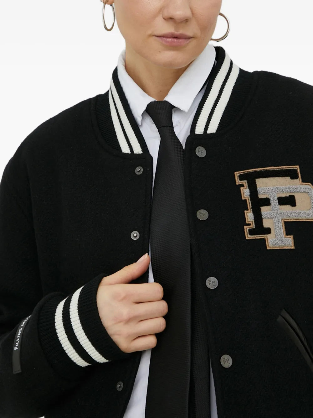 A close-up of a person wearing a black varsity-style jacket with white striped ribbed trim, a large embroidered letter 'F' patch on the chest, and silver buttons, styled over a white collared shirt and black tie.