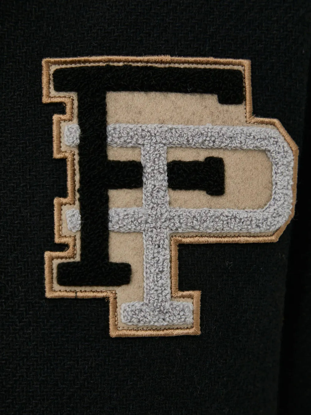 A close-up of a black textured fabric patch featuring a large embroidered letter 'P' in black, beige, and gray.