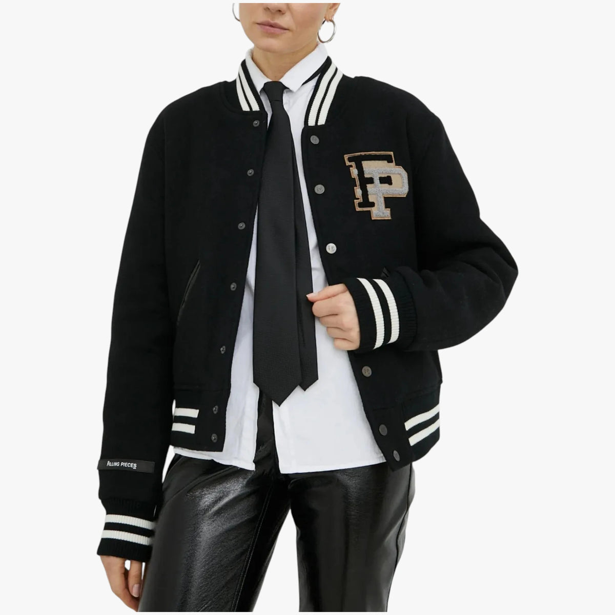 A model wears a black varsity jacket with white and black striped ribbed trim, featuring a gold and white embroidered 'P' logo on the chest. The jacket is styled over a white button-down shirt, a black tie, and black leather pants against a light gray background.