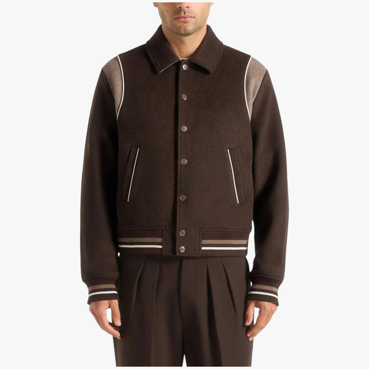 A man wearing a dark brown wool-blend varsity jacket with contrasting light brown shoulder panels and striped ribbed trim, paired with matching dark brown pleated trousers, against a plain light gray background.