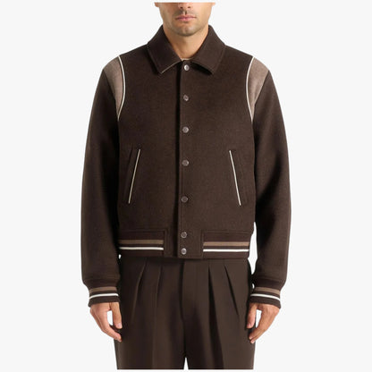 A man wearing a dark brown wool-blend varsity jacket with contrasting light brown shoulder panels and striped ribbed trim, paired with matching dark brown pleated trousers, against a plain light gray background.