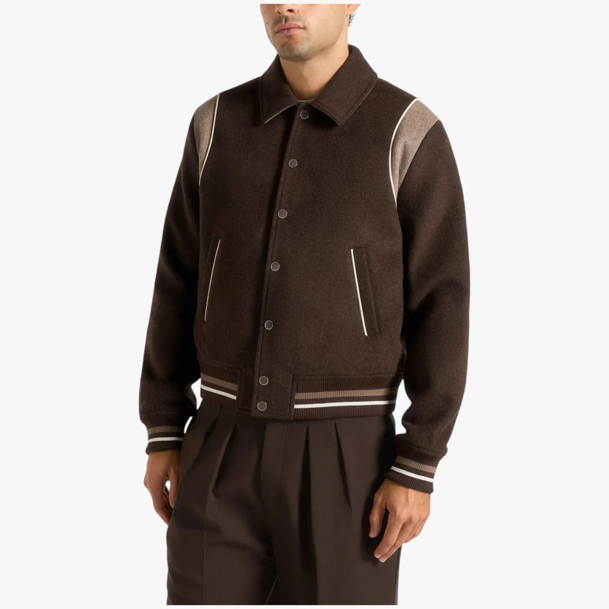 A man wearing a dark brown wool-blend varsity jacket with contrasting light brown shoulder panels and striped ribbing, paired with matching dark brown trousers, against a plain light gray background.