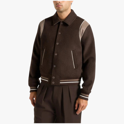 A man wearing a dark brown wool-blend varsity jacket with contrasting light brown shoulder panels and striped ribbing, paired with matching dark brown trousers, against a plain light gray background.