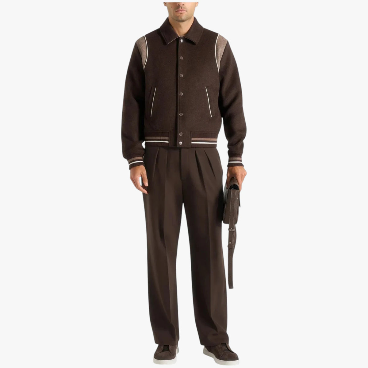 A man models a dark brown varsity-style jacket with contrasting light brown shoulder panels and striped ribbing, paired with matching dark brown trousers and holding a small dark accessory against a plain light gray background.