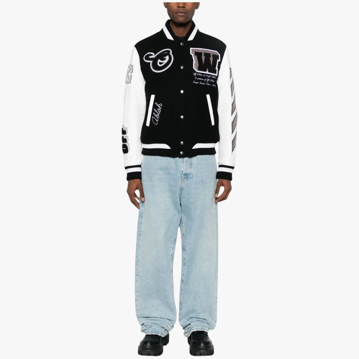 A model wearing a black and white varsity-style bomber jacket with various embroidered patches, paired with light-wash blue jeans and black shoes, shot against a plain white background.