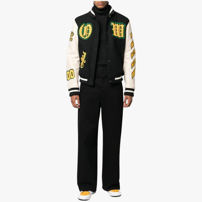 A model wearing a black and cream varsity jacket with green and yellow embroidered patches, paired with black trousers and yellow-and-white sneakers, shot against a plain white background.