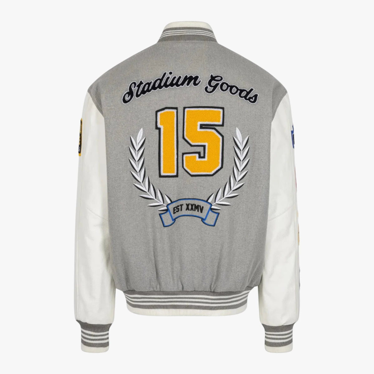 The back of a gray and white varsity-style jacket with 'Stadium Goods' embroidered across the shoulders, a large yellow number 15, and a silver laurel wreath on a light gray background.