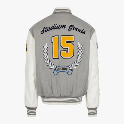 The back of a gray and white varsity-style jacket with 'Stadium Goods' embroidered across the shoulders, a large yellow number 15, and a silver laurel wreath on a light gray background.