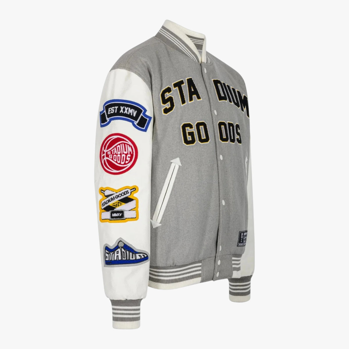 A gray and white varsity-style jacket with multiple colorful embroidered patches on the sleeves and 'STADIUM GO ODS' in black and gold lettering on the chest, shown on a light gray background.