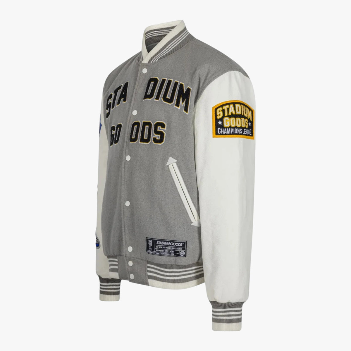 A gray and white varsity-style jacket with 'STADIUM GOODS' lettering on the chest and a 'Stadium Goods Champions 2024' patch on the sleeve, shown on a light gray background.