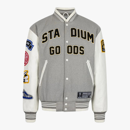 A gray and white varsity-style jacket with 'STADIUM GO ODS' embroidered on the chest, featuring white sleeves with multiple colorful patches, a gray ribbed collar, cuffs, and hem, and white snap-button front closure.